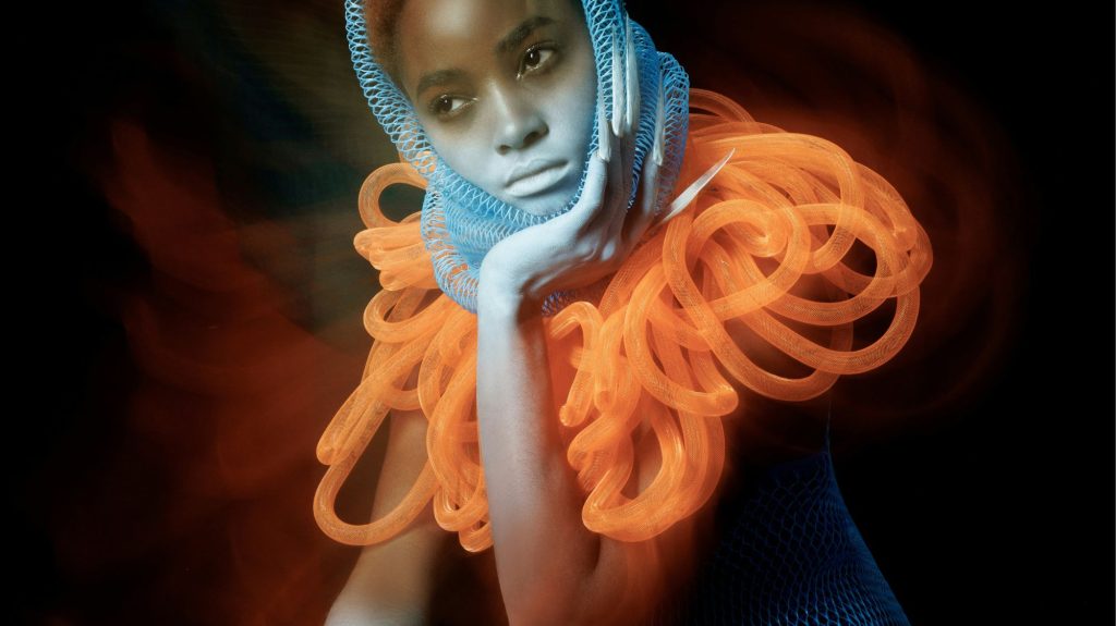 Model in avant-garde fashion featuring vivid orange tubing and blue mesh headpiece, symbolizing AI-influenced creativity and post-human aesthetics.