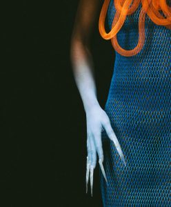 Futuristic fashion close-up featuring a blue mesh dress, elongated sculptural nails, and orange tubing — symbolizing AI’s influence on creativity and identity.