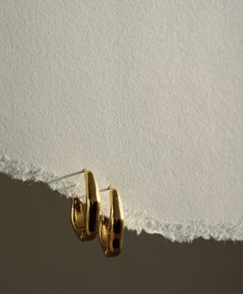 Minimalist gold hoop earrings on torn textured paper, symbolizing refined design and sustainable elegance for Autumn Winter 2026 2027.