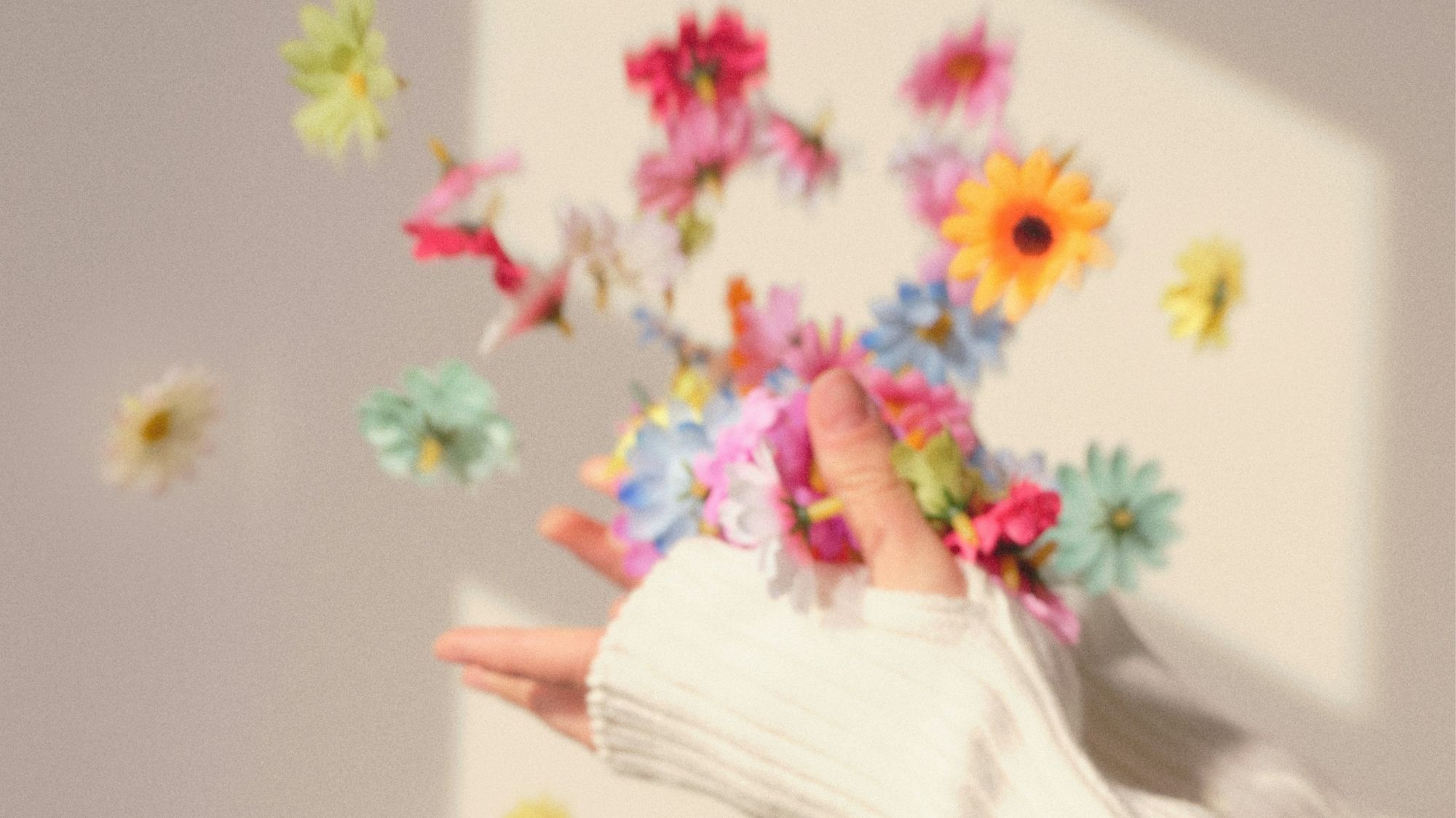 Hands in a white sweater releasing colorful artificial flowers in soft sunlight, symbolizing joy, creativity, and emotional storytelling in fashion trend forecasting.