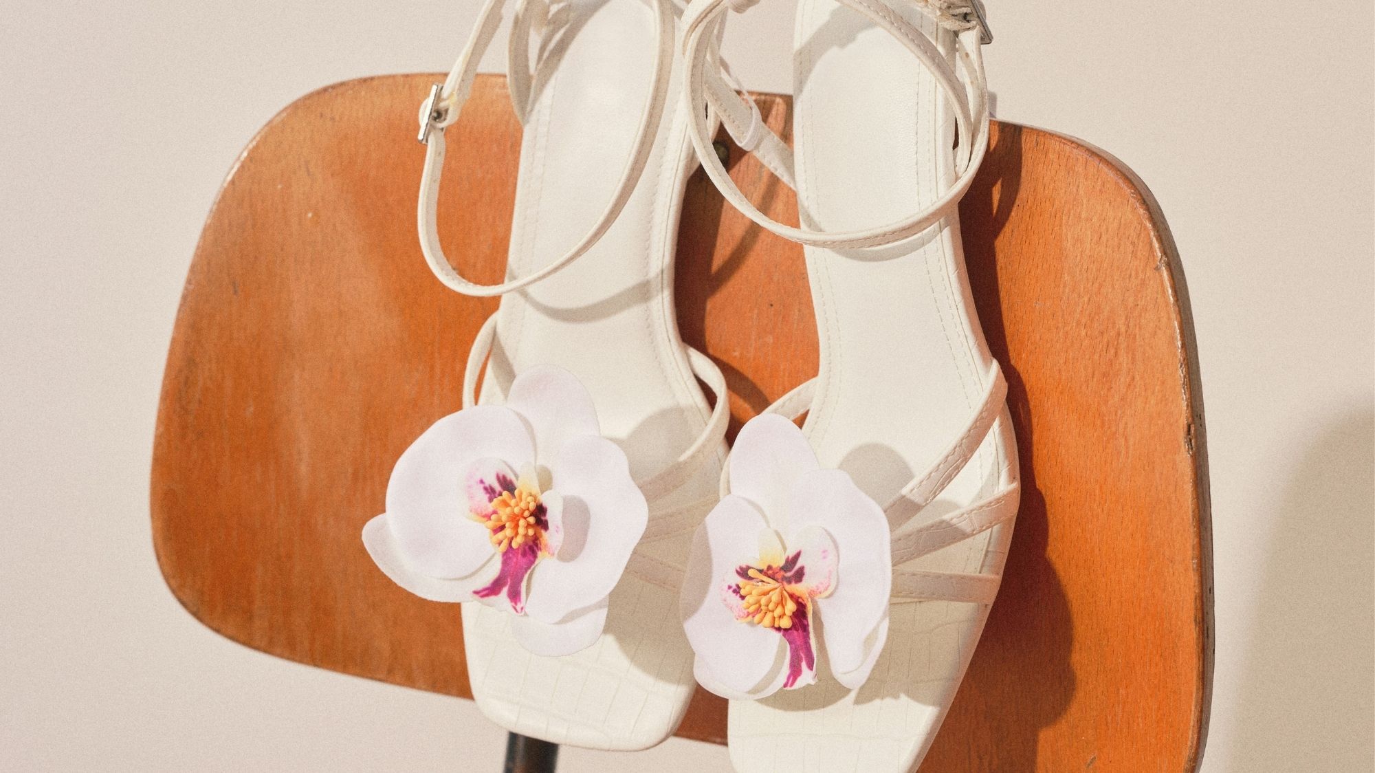 Pair of white sandals with floral embellishments resting on a wooden chair, showcasing romantic detailing and fashion trend forecasting insights.