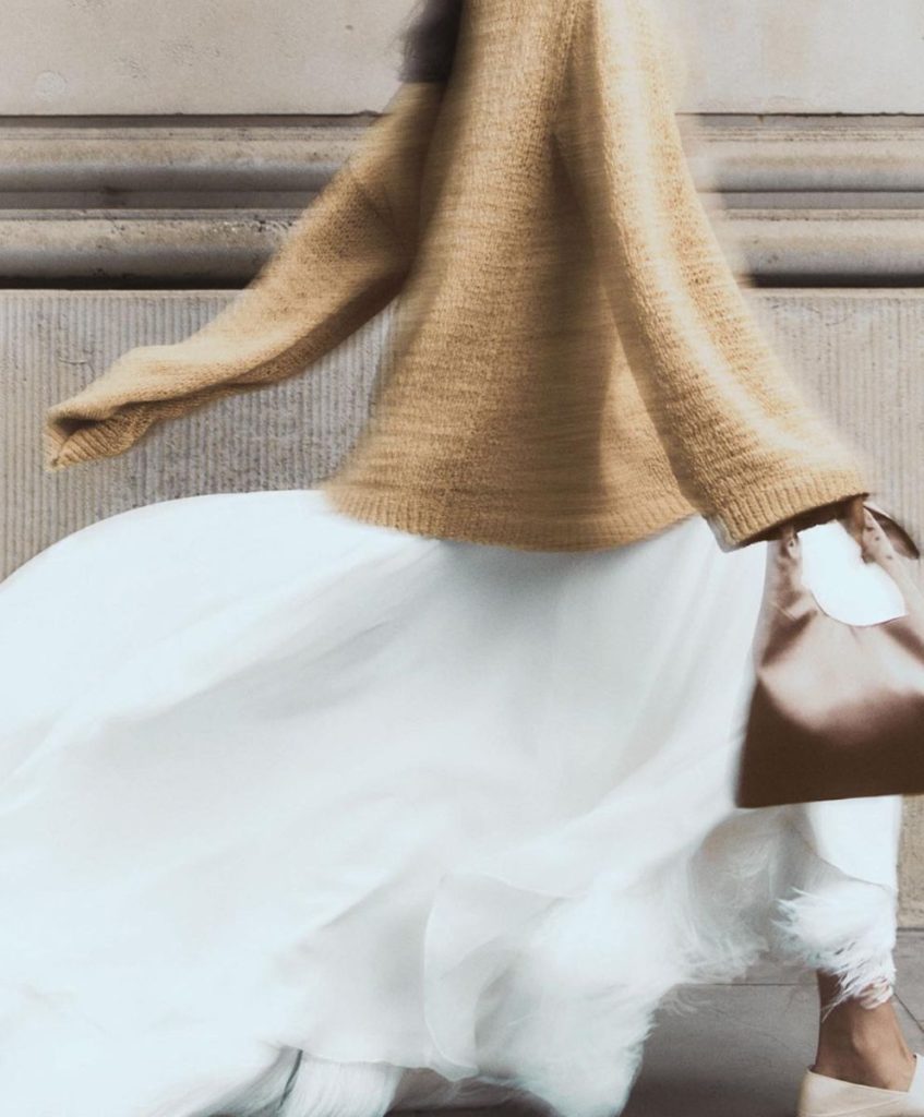 Woman in camel knit sweater and flowing white skirt walking with movement — symbolizing quiet elegance and emotional clarity in fashion.