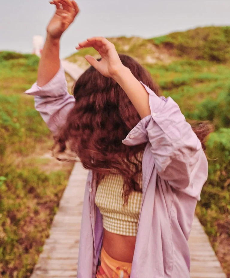 Young woman in summer outfit dancing outdoors on a wooden path, wearing a lilac shirt and gingham crop top.