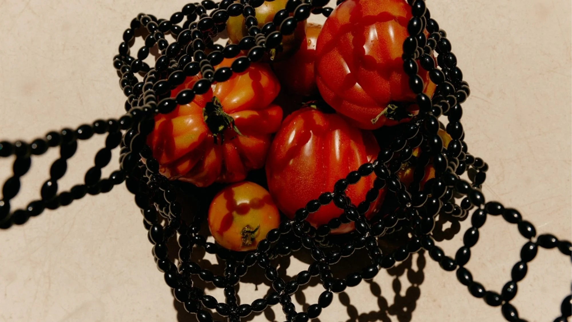 Cluster of red tomatoes entangled in glossy black beaded chains, symbolizing the tension between organic culture and constructed aesthetics, curated by a trend forecasting agency.