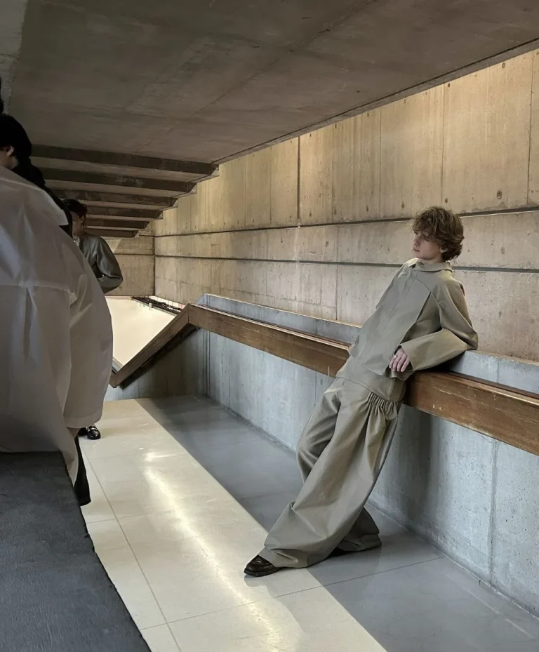 Model in oversized neutral-toned fashion leaning against concrete architecture, reflecting minimalism and cultural repair in Autumn Winter 2027 2028 trend forecasting by C2 Fashion Studio, a leading trend forecasting agency.