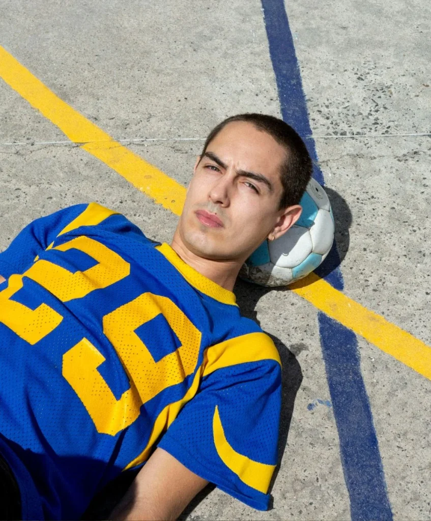 Man in a blue and yellow sports jersey lying on a concrete court with a football behind his head, symbolizing the shift from fast micro-trends to deeper cultural vibes, curated by a trend forecasting agency.