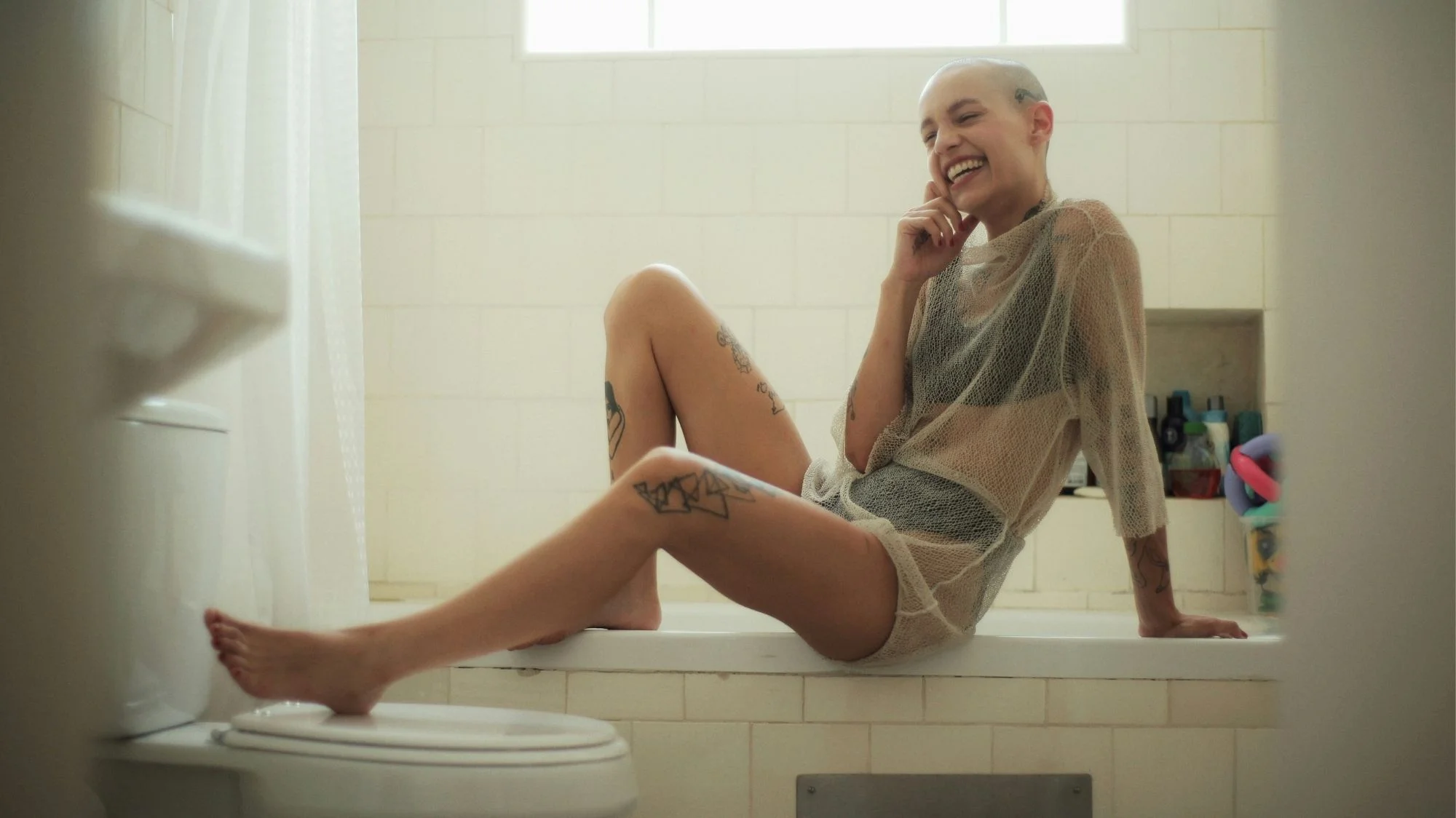 Smiling model with tattoos sitting casually on a bathtub in sheer layered fashion, symbolizing authenticity and imperfection in luxury, curated by C2 Fashion Studio, a global trend forecasting agency specializing in fashion trend forecasting.