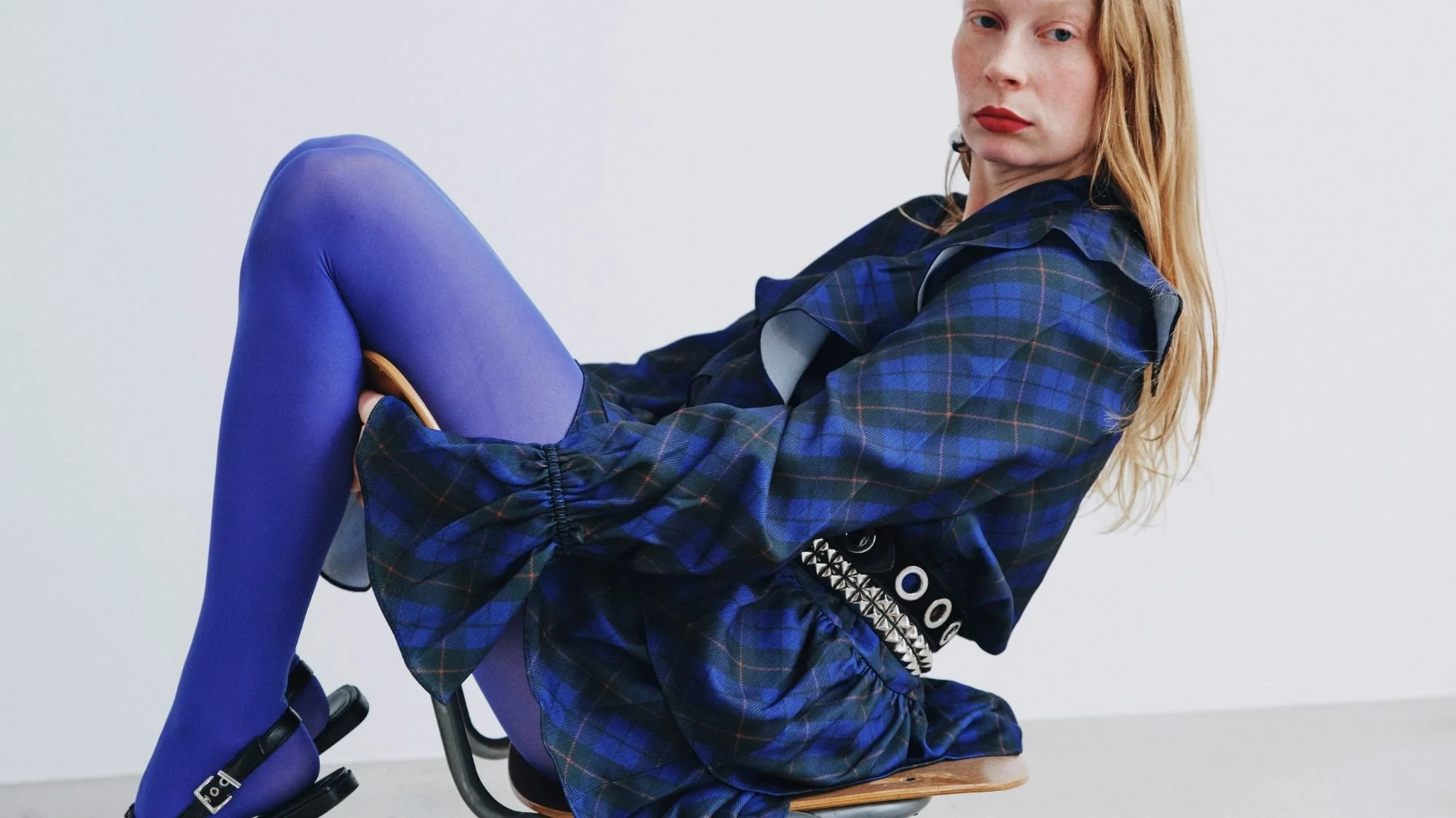 Model in bold blue plaid dress with vibrant tights and studded belt, styled to reflect avant-garde design directions from C2 Fashion Studio, a trend forecasting agency delivering expert fashion trend solutions.