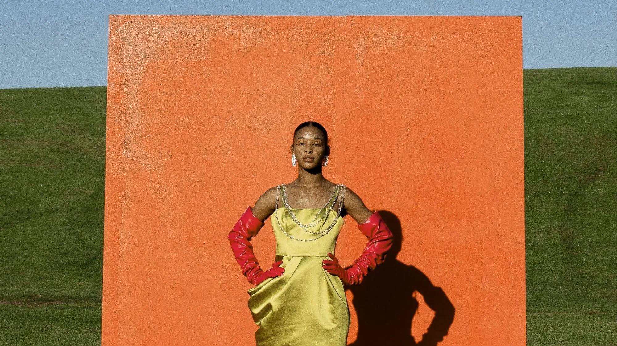 Model in a vibrant yellow dress with red gloves stands before an orange backdrop, symbolizing bold aesthetics and craftsmanship in a fashion trend decoded by C2 Fashion Studio, a leading trend forecasting agency.