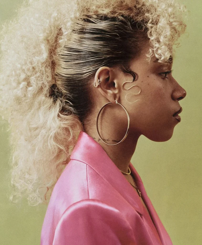 Profile of a model with voluminous curly hair, oversized hoop earrings, and a bold pink blazer, reflecting fragmented styles and cultural acceleration in fashion. Curated by C2 Fashion Studio, a leading trend forecasting agency specializing in fashion trend forecasting.