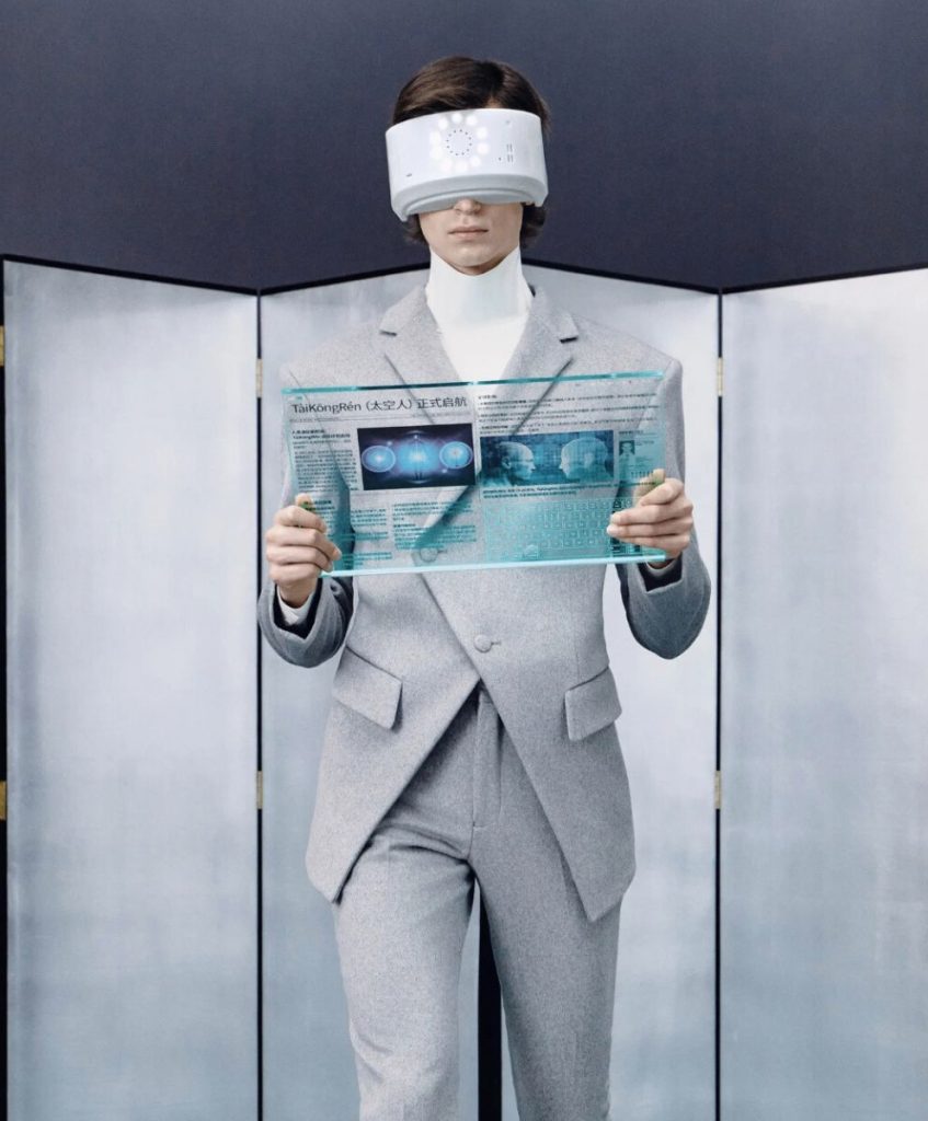 A model in a futuristic grey suit with VR visor and digital tablet, symbolizing the Meta-Executive fashion trend for AW 2026/2027 explored by C2 Fashion Studio, a leading trend forecasting agency.