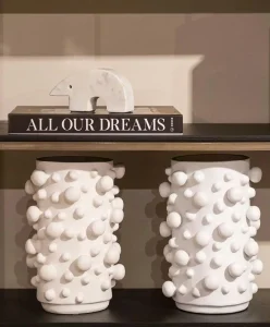 White sculptural ceramic vases with rounded 3D textures displayed on a modern shelf with a marble bear figurine and a book titled “All Our Dreams,” showcasing luxury home decor and contemporary interior design trends.
