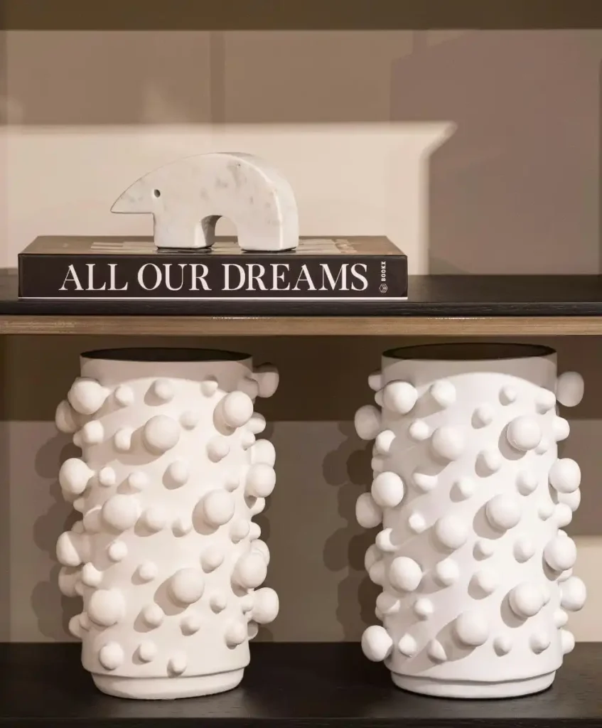 White sculptural ceramic vases with rounded 3D textures displayed on a modern shelf with a marble bear figurine and a book titled “All Our Dreams,” showcasing luxury home decor and contemporary interior design trends.