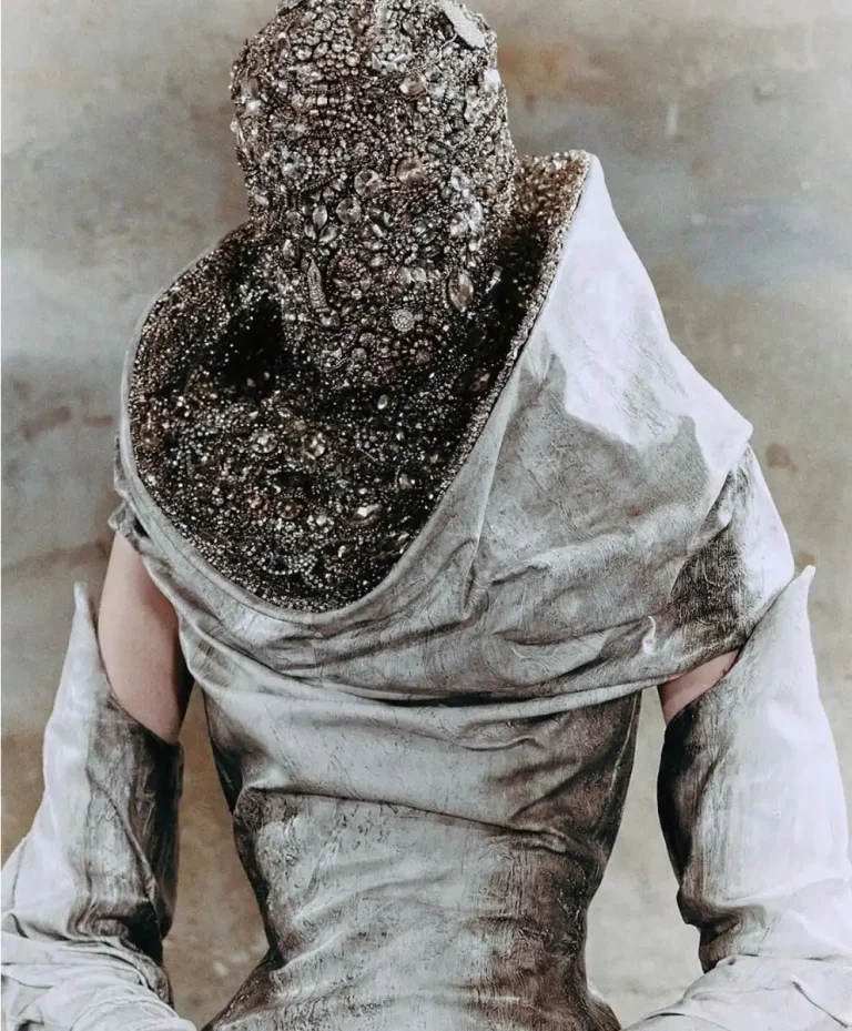 Back view of a model in a distressed silver gown with a heavily encrusted, jewel‑covered headpiece, expressing Entropism and material decay as a fashion trend.