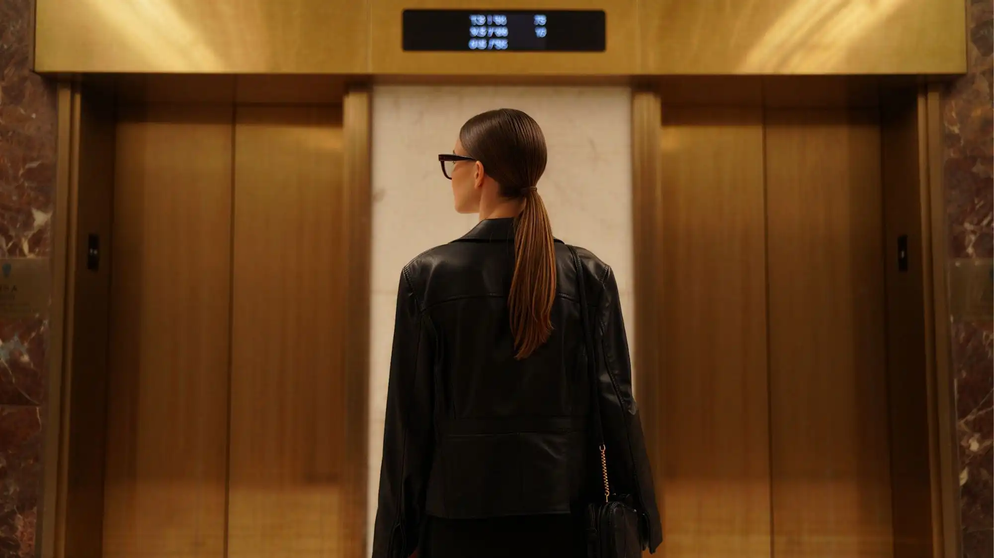 Future Consumer 2028 luxury lifestyle aesthetic featuring a woman in minimalist black leather tailoring standing before golden elevator doors, representing ambition, professional identity, and contemporary luxury culture.