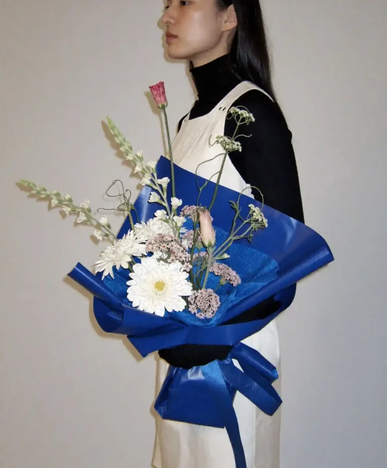 Minimal fashion portrait featuring a woman holding a blue wrapped floral bouquet, representing emotional storytelling, sensory aesthetics, and Future Consumer lifestyle trends for Spring Summer 2026 by C2 Fashion Studio.