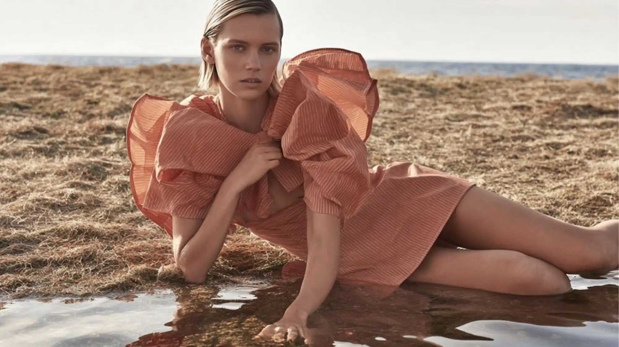 Luminous Balance Spring Summer 2028 Color Trend Forecast featuring a model in a soft coral-pink voluminous dress beside reflective water and sunlit natural landscapes. The image expresses balanced elegance, luminous warmth, emotional optimism, and contemporary femininity through C2 Fashion Studio Trend Forecasting, Trend Intelligence, and Consumer Behavior research.