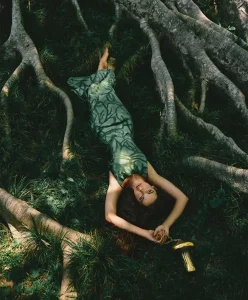 Symbiotic Futures fashion trend for Spring Summer 2027 featuring a model resting among exposed tree roots in a forest landscape, wearing a nature-inspired green dress that reflects biomorphic design, regenerative aesthetics, and bio-intelligent Fashion Trend Forecasting.