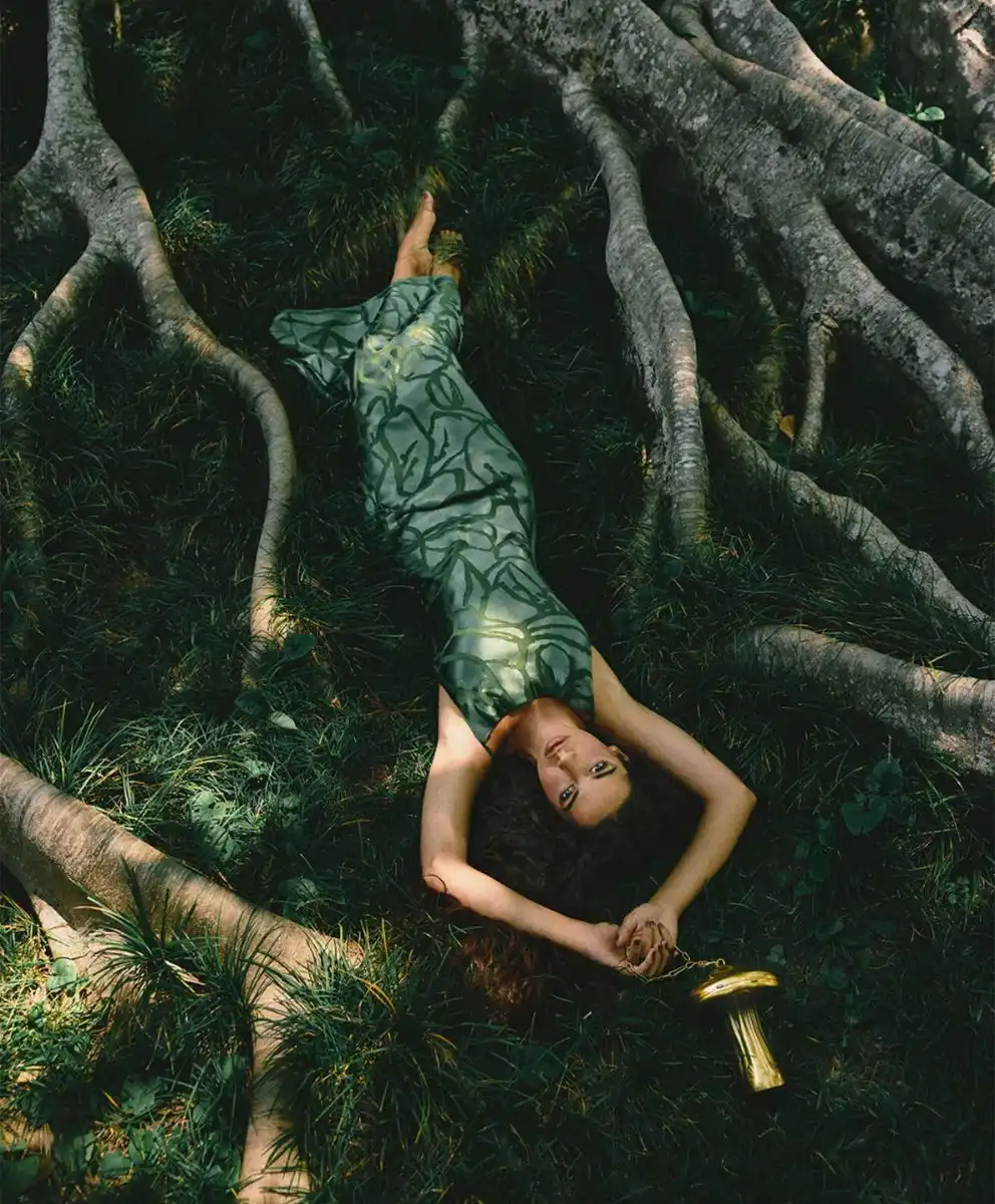 Symbiotic Futures fashion trend for Spring Summer 2027 featuring a model resting among exposed tree roots in a forest landscape, wearing a nature-inspired green dress that reflects biomorphic design, regenerative aesthetics, and bio-intelligent Fashion Trend Forecasting.